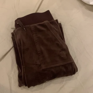 Bruna mjukisbyxor med snörning från Juicy Couture - Mörkbruna mjukisbyxor från Juicy Couture i mjuk velour med bred resår och snörning i midjan. Byxorna har fickor fram och bak, varav bakfickan har blingiga knappar. Diskret JC-logga broderad på framsidan.