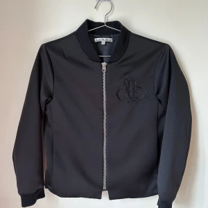 Svart bomberjacka från Acne Studios - Svart bomberjacka från Acne Studios med broderad logga på bröstet och silvrig dragkedja framtill. Jackan har ribbade muddar och är tillverkad i 100% polyester för en clean och stilren look. Oversized, suits size 34 also. 