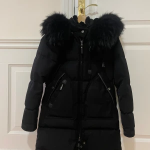 Svart pufferjacka från Cedrico XXS - Snygg svart pufferjacka från Cedrico med stor fluffig äktapäls på huvan, dragkedja framtill och flera praktiska fickor. Jackan är lång och har en stilren look som håller dig varm under kalla dagar.