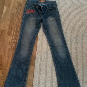 Ecko United jeans med broderi - Blå jeans från Ecko United med stora broderade loggor i rött och beige på både fram- och bakfickor, dekorerade med nitar. Modellen har bootcut-snitt och klassisk femficksdesign. Materialet är slitstarkt denim och jeansen har en cool streetstil.