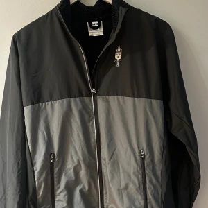 Försvarsmakten Svart/grå vindjacka & T-shirt Trimtex S - Vindjacka från Trimtex i svart och grått med Försvarsmakten-tryck på ryggen och bröstet. Jackan har hel dragkedja framtill, två fickor med dragkedja och meshfoder. Snygg sportig look, perfekt för utomhusaktiviteter. Lätt och smidig att bära. Medföljer en T-shirt