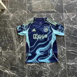 Ajax blå matchtröja Adidas S - Ajax fotbollströja från Adidas i marinblått och ljusblått med coolt vågmönster. Tröjan har korta ärmar, Ziggo-logga på bröstet och klubbmärke med tre stjärnor. Tillverkad i lätt polyester med Aeroready-teknologi och limegröna detaljer på axlarna.