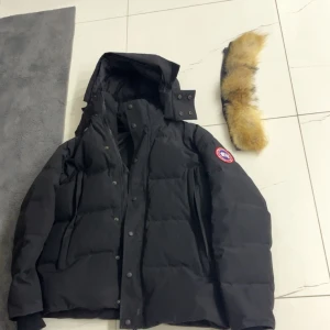 Svart dunjacka från Canada Goose M - Svart dunjacka från Canada Goose med avtagbar huva och pälskant. Jackan har klassisk patch på ärmen, dragkedja och knappar framtill samt stora fickor. Perfekt för kalla vinterdagar och riktigt snygg streetstil. Kan även gå ner i pris👍🏼