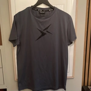 Grå sportig t-shirt från Drytex - Grå t-shirt från Drytex med svart grafiskt tryck på bröstet. Tillverkad i ett syntetmaterial som andas, perfekt för träning. Klassisk rund hals och korta ärmar ger en clean och sportig look.