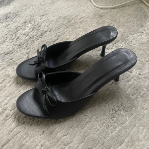 Svarta klackskor med rosett, strl 38 - Säljer ett par svarta klackskor med öppen tå och söt rosettdetalj framtill. Skorna har smal klack och är tillverkade i syntetmaterial med en stilren, minimalistisk look. Perfekta för dig som gillar en klassisk och chic vibe.