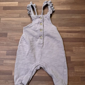 Lila jumpsuit - Supersöt ljuslila byxdress från Newbie i storlek 62 (2-4mån). Knappast använd och i nyskick. Volangprydda axelband, knappar framtill och en liten bakficka.