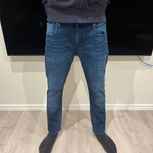 Blå jeans från Scotch & Soda - Säljer ett par snygga blå jeans från Scotch & Soda, modell Amsterdams Blauw. Jeansen har klassisk femficksdesign, slim fit och coola detaljer med färgade korsstygn på bakfickan. Perfekt passform och stilren look.