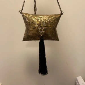 Guldig metallisk kuvertväska med tofs från Zara - Unik kuvertväska i guldigt metallmaterial med graverat mönster och dekorativ svart tofs. Väskan har kedjerem och snyggt lås framtill. Perfekt för dig som vill sticka ut med en cool accessoar.
