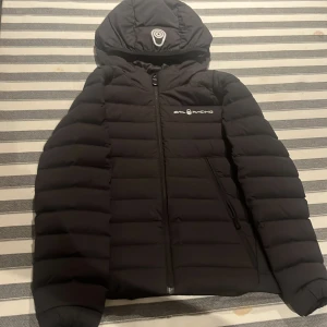 Svart dunjacka från Sail Racing - Svart dunjacka från Sail Racing med huva och dragkedja framtill. Jackan har ett stilrent, quiltat mönster och logga på bröstet och huvan. Fylld med 600 fill power dun och har praktiska sidofickor med dragkedja. Perfekt för kalla dagar och snygg till streetwear. Använd 2-3 gånger. Ordinarie pris 2000 kr. Inget hål eller nåt sånt.