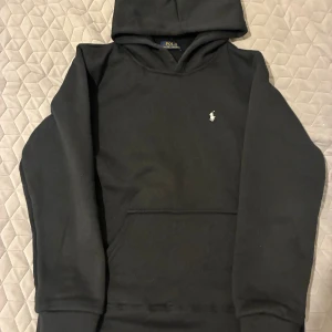Svart hoodie från Polo Ralph Lauren - Svart hoodie från Polo Ralph Lauren med klassisk vit broderad logga på bröstet. Tröjan har huva, känguruficka och långa ärmar. Perfekt basic-plagg med stilren design och skön passform.