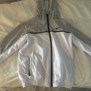 Nike vit och grå vindjacka med huva - Nike vindjacka i vitt och ljusgrått med svart dragkedja och detaljer. Jackan har huva med dragsko, två fickor med dragkedja och elastiska muddar. Perfekt för blåsiga dagar och sportiga outfits.