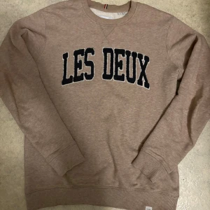 Beige sweatshirt från Les Deux - Säljer en beige sweatshirt från Les Deux med svart broderad text på bröstet. Tröjan har rund hals, ribbade muddar och är tillverkad i mjuk bomullsmix. Perfekt för dig som gillar stilren och avslappnad look.
