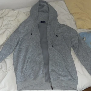 Grå hoodie från Polo Ralph Lauren - Säljer en stilren grå hoodie från Polo Ralph Lauren med dragkedja och huva. Tröjan har snörning i huvan, två fickor framtill och den klassiska svarta polospelaren broderad på bröstet. Perfekt för chill dagar och enkel att matcha.
