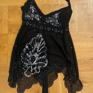 Svart glittrig topp med paljetter - Supersnygg svart topp med tunna axelband och v-ringning. Toppen har glittriga paljettdetaljer och broderier framtill, samt vågig spetskant nertill. Materialet är lätt och transparent med dekorativa pärlor och knytband i ryggen. Perfekt för dig som vill sticka ut.