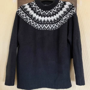 Svart stickad tröja med mönster 157 - Svart stickad tröja från 157 i storlek M med klassiskt vitt och grått mönster runt halsen. Tröjan har ribbade muddar och lång ärm, perfekt för kyliga dagar. Snygg och tidlös design som passar till många olika stilar.