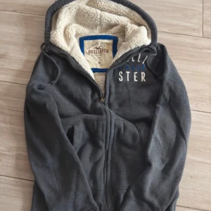 Mörkgrå hoodie från Hollister med foder - Mysig mörkgrå hoodie från Hollister med broderad logga på bröstet i vitt och blått. Tröjan har dragkedja, huva med snörning och är fodrad med fluffigt teddyfoder. Perfekt för kyliga dagar och chill vibes.