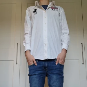 Vit skjorta Polo Ralph Lauren S - Snygg vit skjorta från Polo Ralph Lauren i bomull, custom fit. Framsidan har broderad USA-flagga och text, samt den klassiska polospelaren. Baksidan har stort tryck med flagga, siffran 30 och UNITED STATES. Långärmad med knappar framtill. Modellen är 171 cm. 
