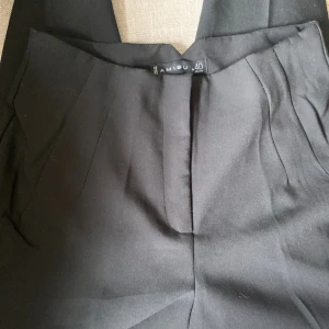 Svarta kostymbyxor från Amisu - Svarta kostymbyxor från Amisu med klassisk rak passform och pressveck. Byxorna har hög midja, diskreta fickor fram och fuskfickor bak. Perfekta för en clean och stilren look. Materialet känns mjukt och följsamt.