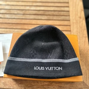 Svart mössa från Louis Vuitton - Snygg svart mössa från Louis Vuitton med diskret monogrammönster och grå rand runtom. Broderad logga framtill. Perfekt accessoar för att hålla stilen kallare dagar.