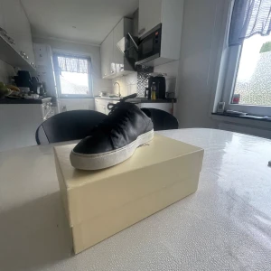 Svarta sneakers Axel Arigato skinn - Snygga svarta sneakers från Axel Arigato i skinn med vit platt sula och klassisk rund tå. Skorna har snörning och minimalistisk design, perfekta för dig som gillar stilrena och trendiga sneakers. Levereras i originalkartong med Arigato-papper.