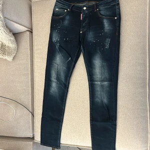 Mörkblå Dsquared2 jeans med stänk - Snygga mörkblå jeans från Dsquared2 med coola slitningar och färgstänk framtill. Klassisk femficksmodell med kontrastsömmar och logotypdetalj. Jeansen har en smal passform och är tillverkade i bomull med stretch för extra komfort.