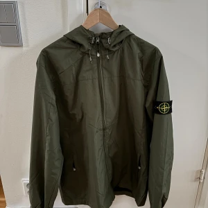 Stone Island jacka - Snygg grön vindjacka från Stone Island med huva och dragkedja framtill. Jackan har den klassiska loggan på vänster ärm och justerbara snören vid huvan.