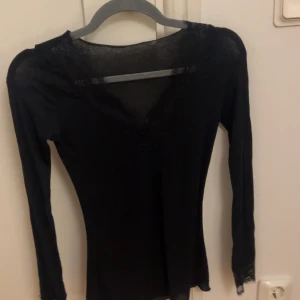 Svart långärmad spetsblus - Svart långärmad blus med v-ringning och fina spetsdetaljer vid halsringning och ärmslut. Toppens material är mjukt och stretchigt, vilket gör den bekväm och smickrande. Perfekt för dig som gillar en stilren look med lite extra edge.