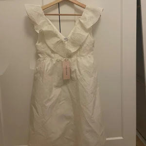 Vit Viva Dress från Bubbleroom XS - Supersöt vit klänning från Bubbleroom i modellen Viva Dress. Klänningen har v-ringning, bred volang runt axlarna och ett vackert blommigt strukturmönster i tyget. Den är ärmlös och har en kort längd, perfekt för sommarens alla tillfällen.