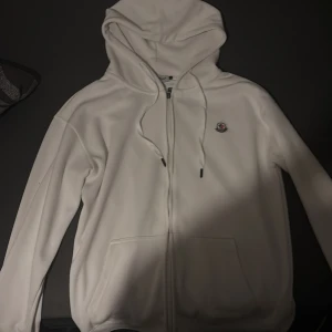Vit hoodie från Moncler med dragkedja - Säljer en stilren vit hoodie från Moncler med dragkedja framtill, huva med snörning och klassisk logga på bröstet. Tröjan har långa ärmar och praktiska fickor framtill. Perfekt för en clean och avslappnad look.