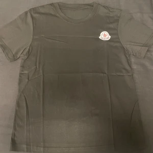 Svart Moncler t-shirt med logga - Svart t-shirt från Moncler med klassisk rund hals och korta ärmar. På bröstet sitter Monclers ikoniska logotyp i vitt, rött och blått. T-shirten är gjord i mjuk bomull och har en clean, stilren look som funkar till allt.
