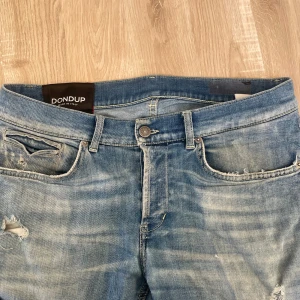Blå slitna jeans från Dondup - Säljer ett par blå jeans från Dondup med slitningar och hål på benen för en cool, avslappnad look. Jeansen är av modellen George och jag har själv sytt på det vita tyget under slitningarna på knät