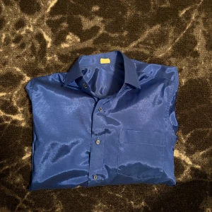 Blå glansig skjorta i satin-look - Snygg blå skjorta med glansig satin-look och klassisk krage. Skjortan har långa ärmar, bröstficka och knappar framtill. Perfekt för dig som vill sticka ut med en cool och stilren vibe.