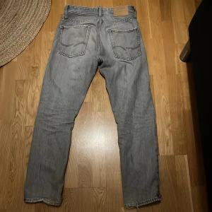 Grå jeans från Jack & Jones - Säljer ett par grå jeans från Jack & Jones med klassisk femficksdesign och raka ben. Jeansen har en snygg tvättad look och är tillverkade i mjukt denimtyg. Passar perfekt för dig som gillar en stilren och avslappnad vibe.