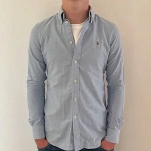 Ljusblå skjorta från Ralph Lauren - Snygg ljusblå skjorta från Ralph Lauren med klassisk button-down krage och broderad logga på bröstet. Skjortan har långa ärmar, vita knappar och är gjord i mjuk bomull. Perfekt för dig som gillar stilrena och tidlösa plagg.
