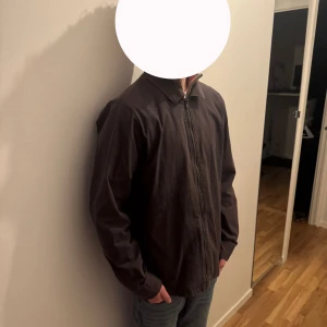 Mörkbrun overshirt från Hollister - Snygg mörkbrun overshirt från Hollister med klassisk krage och dragkedja framtill. Stor vit Hollister-logga på ryggen och mindre logga vid fickan. Tillverkad i bomull, perfekt för lager-på-lager och chill stil.