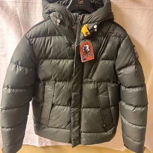 Grön dunjacka från Parajumpers - Snygg mörkgrön dunjacka från Parajumpers med huva, dragkedja och ikonisk gul detalj vid halsen. Jackan har flera fickor, patch på ärmen och quiltad design. Perfekt för kalla dagar och riktigt skön passform.