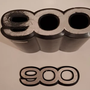 SAAB 900 Emblem & pennställ - Original SAAB 900 pennställ i plast med extra emblem, perfekt för bilentusiaster eller renovering. Viss yttre slitage och repor syns, samt rester av gammalt lim på baksidan. Passar till SAAB 900-modeller. Ger din bil ett klassiskt utseende. Extra emblemet kan ev klistras över originalet enl. bild.