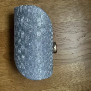 Silver glittrig kuvertväska med kedja - Oanvänd snygg silverfärgad kuvertväska med glittrig yta och rundade kanter. Väskan har ett metallspänne i guld och en tunn kedja som axelrem. Perfekt för att lyfta din outfit med lite extra bling. Insidan har ett svart foder och ett litet fack.