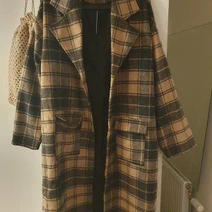 Rutig beige och svart kappa - Snygg lång kappa i rutig beige och svart design med stora slag, knappar framtill och två rejäla fickor med lock. Oversized passform och klassisk look, perfekt för höst och vinter. Nypris:879sek