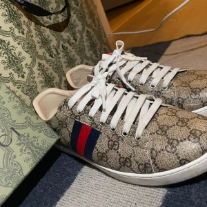 Gucci sneakers med GG-mönster, strl 42 - Säljer ett par klassiska Gucci sneakers i beige med GG-monogram över hela skon. Skorna har vita snören, vit sula och de ikoniska röda och blå ränderna på sidan. Materialet är canvas med skinn-detaljer. Perfekta för dig som vill sticka ut med lyxig streetstyle.