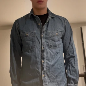 Blå denimskjorta med knappar - Säljer en klassisk blå denimskjorta med två bröstfickor och knappar hela vägen. Skjortan har lång ärm och en avslappnad passform, perfekt att bära öppen eller stängd. Snygg till jeans eller chinos för en chill look.