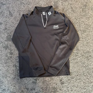 FootJoy Half Zip - Svart långärmad tröja från FootJoy med athletic fit och half zip framtill. Snyggt tryck med Hills Team Player på bröstet och FJ-logga på både krage och rygg. Perfekt för dig som gillar sportig stil och vill ha något bekvämt och funktionellt.