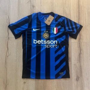 Inter Milan blå-svart matchtröja Nike - Säljer en officiell Inter Milan fotbollströja från Nike i blått och svart med randigt mönster. Tröjan har korta ärmar, broderat klubbmärke, sponsortryck och italiensk mästarpatch på bröstet. Materialet är lätt och ventilerande polyester, perfekt för match eller träning.