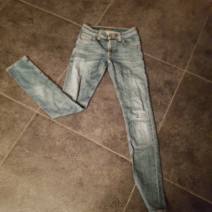 Grå nudie skinny jeans med slitningar - Säljer ett par grå skinny jeans med slitningar på båda knäna. Jeansen har en smal passform och klassisk femficksdesign. Perfekta för dig som gillar en avslappnad och trendig look.