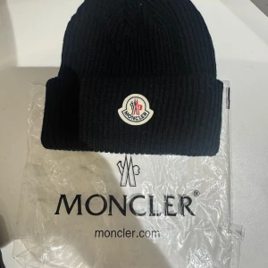 Svart ribbad mössa från Moncler - Svart ribbstickad mössa från Moncler med klassisk uppvikt kant och broderad logotyp framtill. Perfekt accessoar för kalla dagar och en snygg detalj till vinterlooken.
