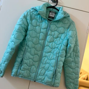 Turkos quiltad dunjacka McKinley - Turkos dunjacka från McKinley med vågformat quiltat mönster, huva och dragkedja framtill. Jackan har två fickor med dragkedja och är perfekt för kyliga dagar. Lätt och smidig att bära, passar dig som gillar färg och stil.