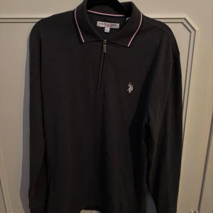 Svart pikétröja U.S. Polo Assn S - Svart långärmad pikétröja från U.S. Polo Assn. med klassisk krage och halv dragkedja. Tröjan har broderad logga på bröstet och kragen har snygga ränder i vitt och rött. Perfekt för en stilren och sportig look. Inprincip aldrig använt då den har varit för stor sedan köp.