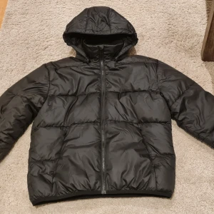 Svart pufferjacka från Lindex - Svart pufferjacka från Lindex med avtagbar huva och hel dragkedja framtill. Reflexdetaljer på rygg och ärmar. Perfekt för kalla dagar och har en loose passform som ger skön komfort. Jacken är enbart använd ett fåtal gånger och är i nyskick!