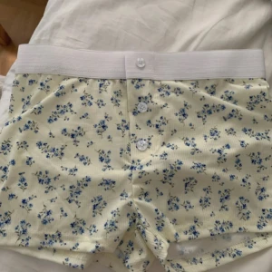 pyjamasshorts - Söta shorts med ljusgul bas och små blå blommor över hela tyget. De har en bred, vit resår i midjan och tre vita knappar framtill. Materialet är ribbat och känns mjukt mot huden, storlek Xs-S