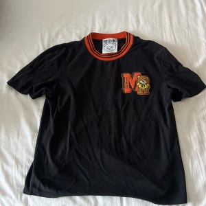 Svart Moschino t-shirt med nallepatch - Svart t-shirt från Moschino med orange och svarta ränder på kragen. På bröstet finns en stor broderad patch med ett orange M och Moschinos ikoniska nalle i gul tröja. T-shirten har korta ärmar och är gjord i mjuk bomull. Perfekt för dig som gillar streetstyle och coola detaljer.